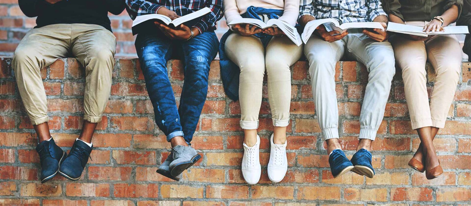 Several students sitting on a brick wall, reading books.