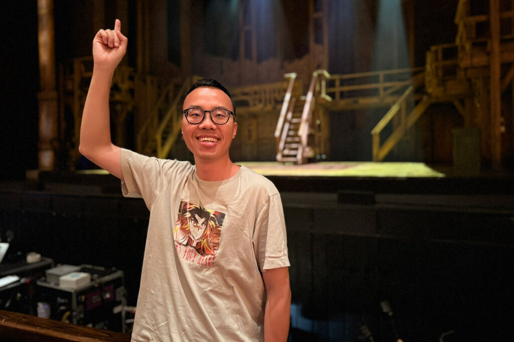 A man with glasses wearing a light coloured t-shirt smiles for the camera while pointing up.