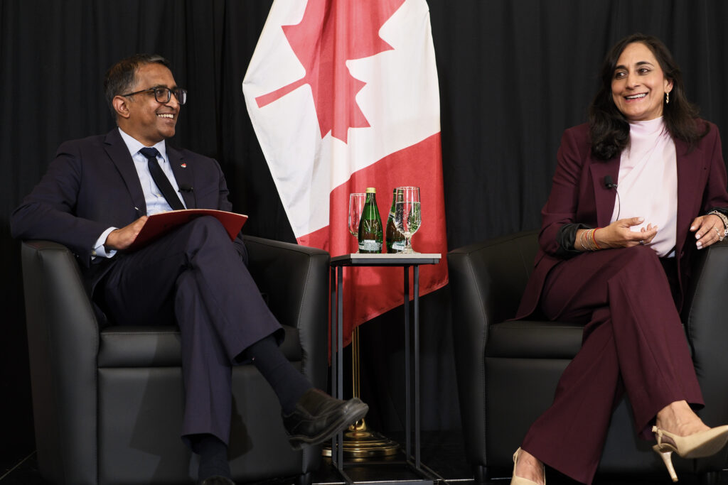 A man and a woman sitting in arm chairs with a black pipe and drape and the Canada flag behind them.