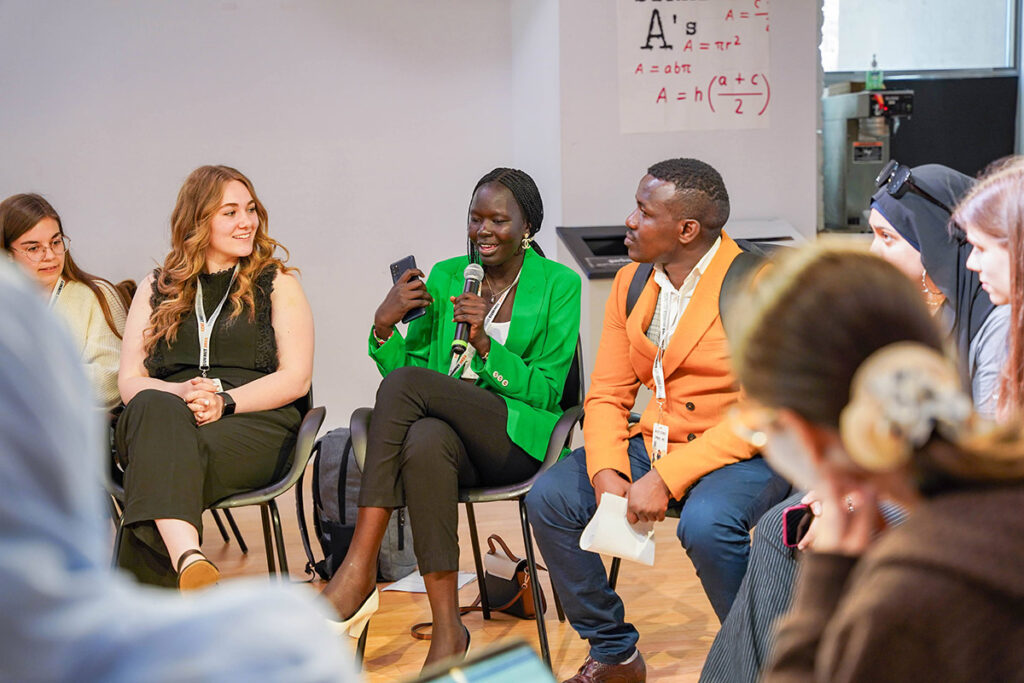 Nyabuol Biel Gang, wearing a green blazer, speaks into a microphone during a group discussion.