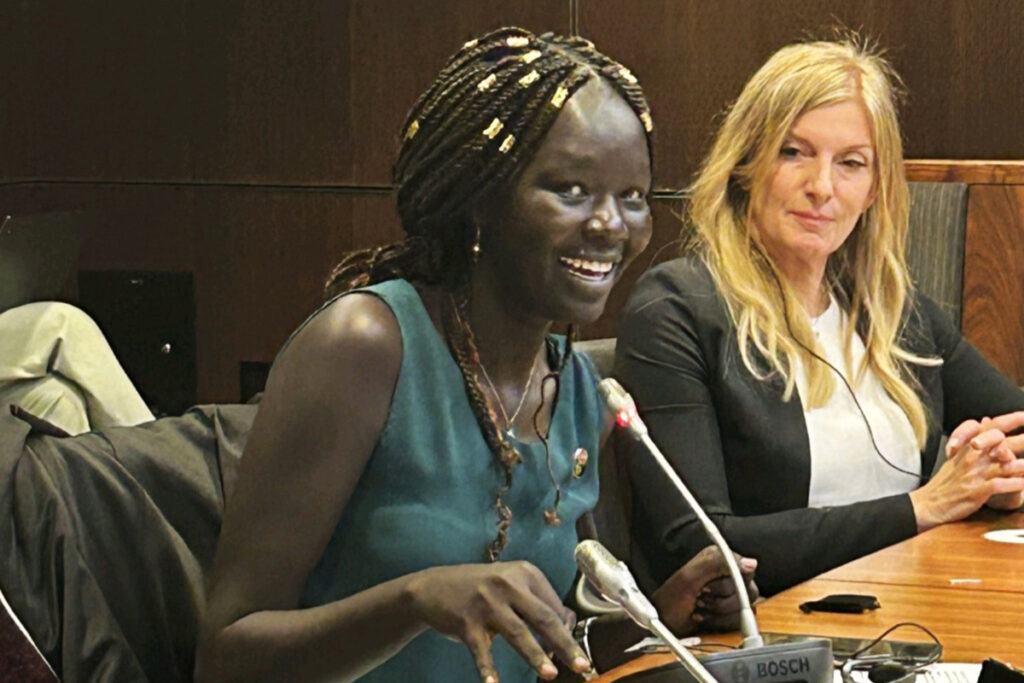 Nyabuol Biel Gang speaks at a table with a microphone while another woman listens.