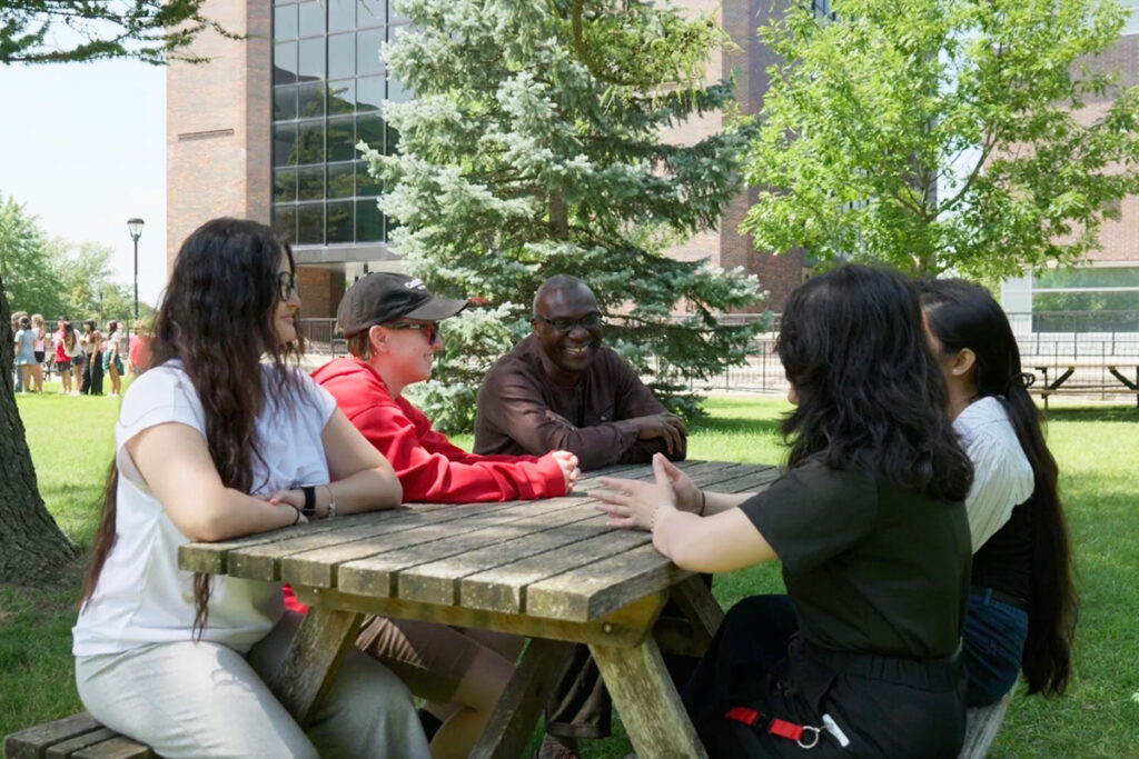 Five people having a discussion at a picnic table surrounded by trees.