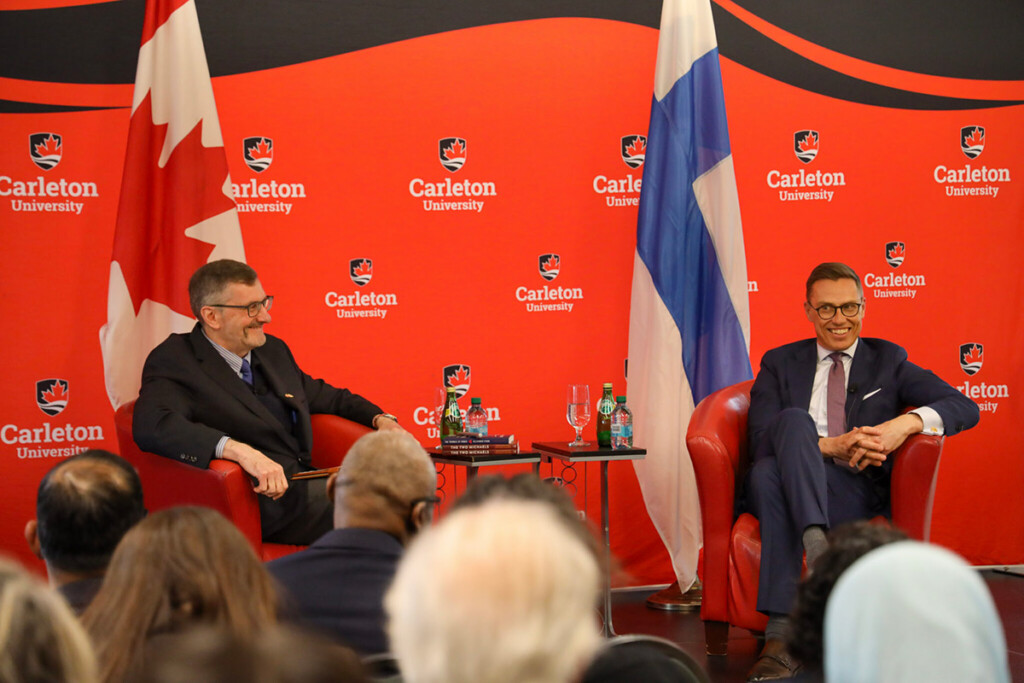 Two people seated on a stage, having a discussion, in front of an audience.