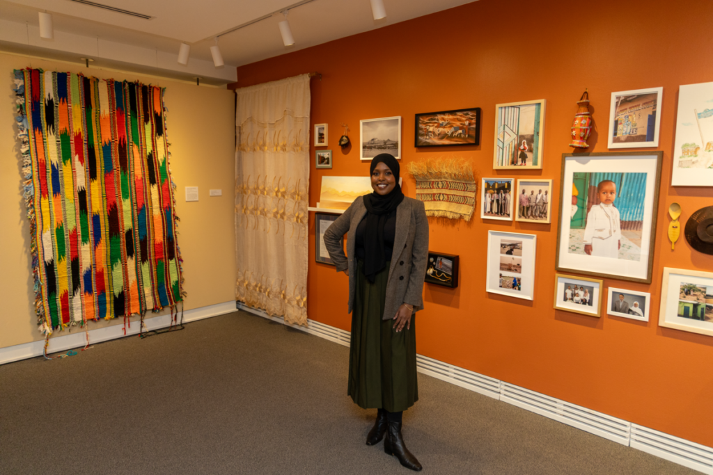 Faisa Omer stands in front, featuring a tapestry to the left wall and portraits on the right wall. 