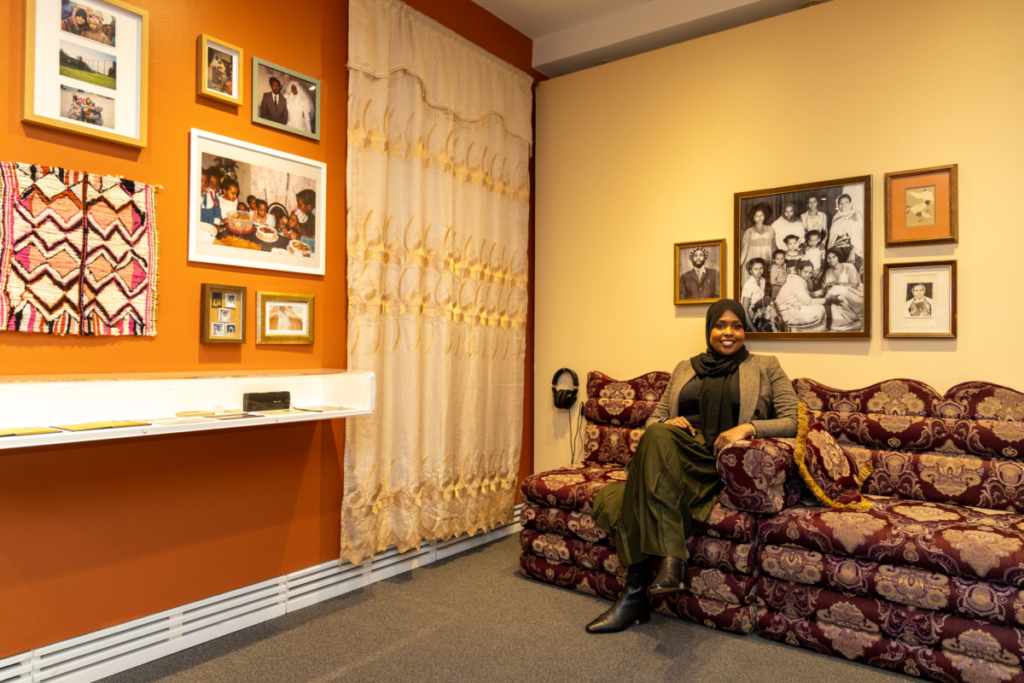 A section of the exhibit, set up like a Somali living room with a traditional couch, with Faisa Omer seated. 