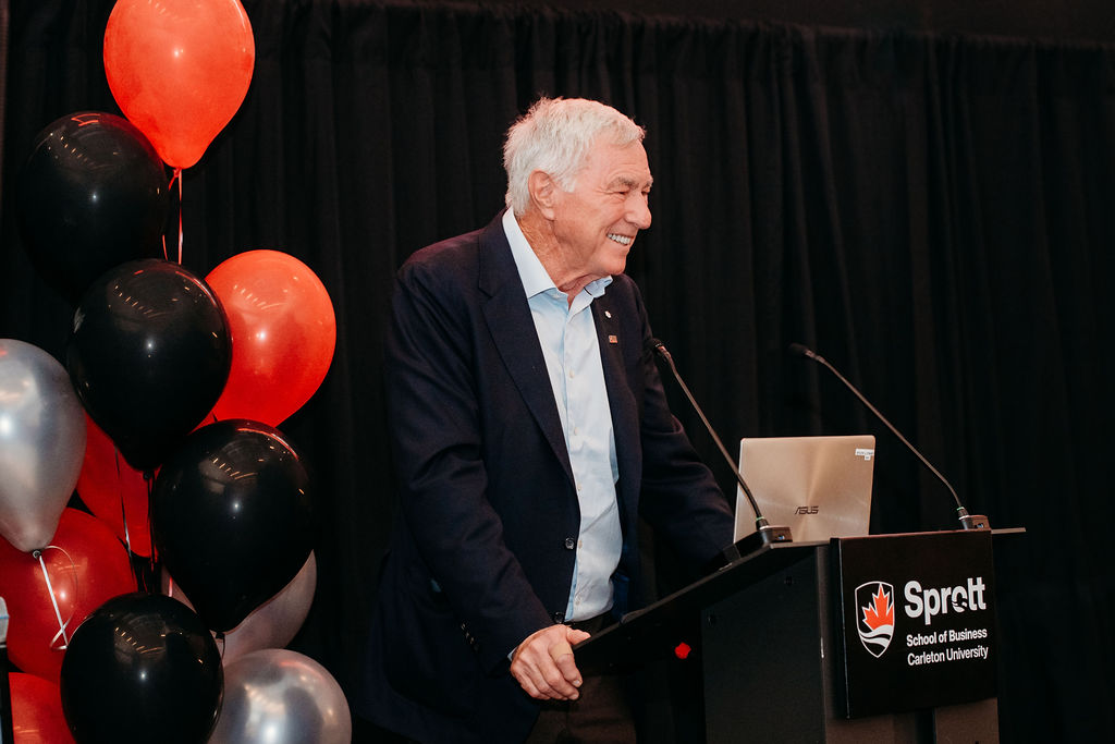 Image of a man standing in front of a podium with a microphone. He is in front of a black background and balloons.