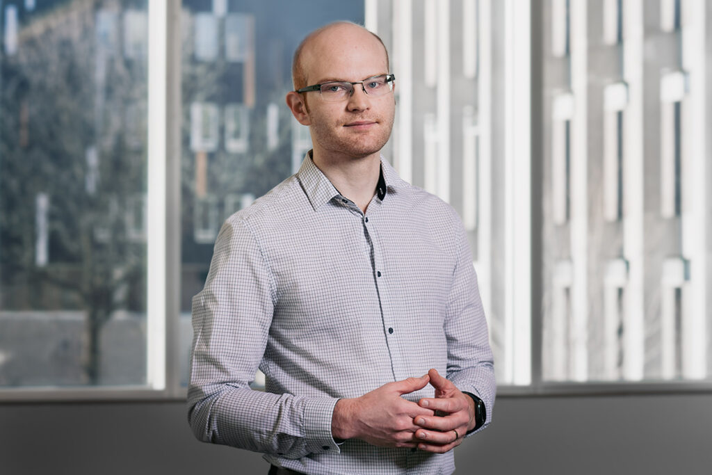 Man wearing glasses and a checked shirt standing by windows.