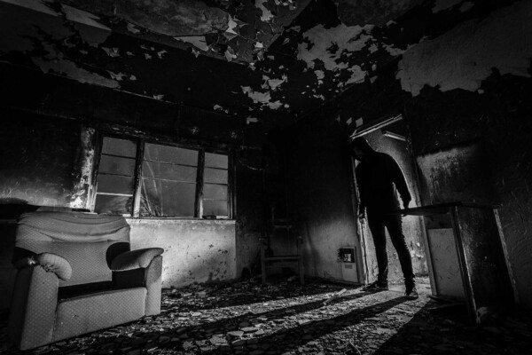 Shadowy figure in the doorway of a dark, abandoned room with peeling paint and a worn armchair.