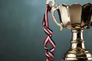 A trophy sits in front of a chalkboard background. A ribbon that would hold a medal is tied to the trophy's arm.