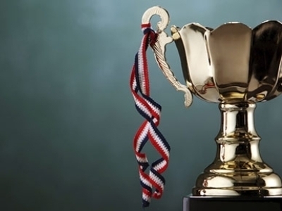 A trophy sits in front of a chalkboard background. A ribbon that would hold a medal is tied to the trophy's arm.