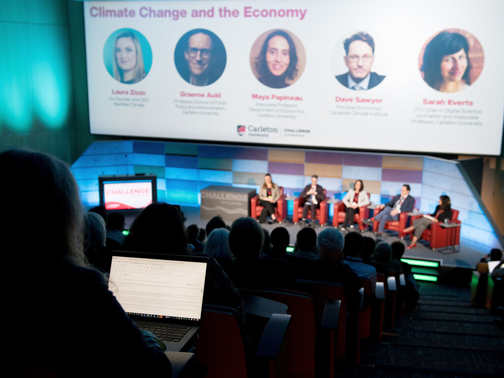 A from behind view of a crowd watching a panel of speakers at a conference.
