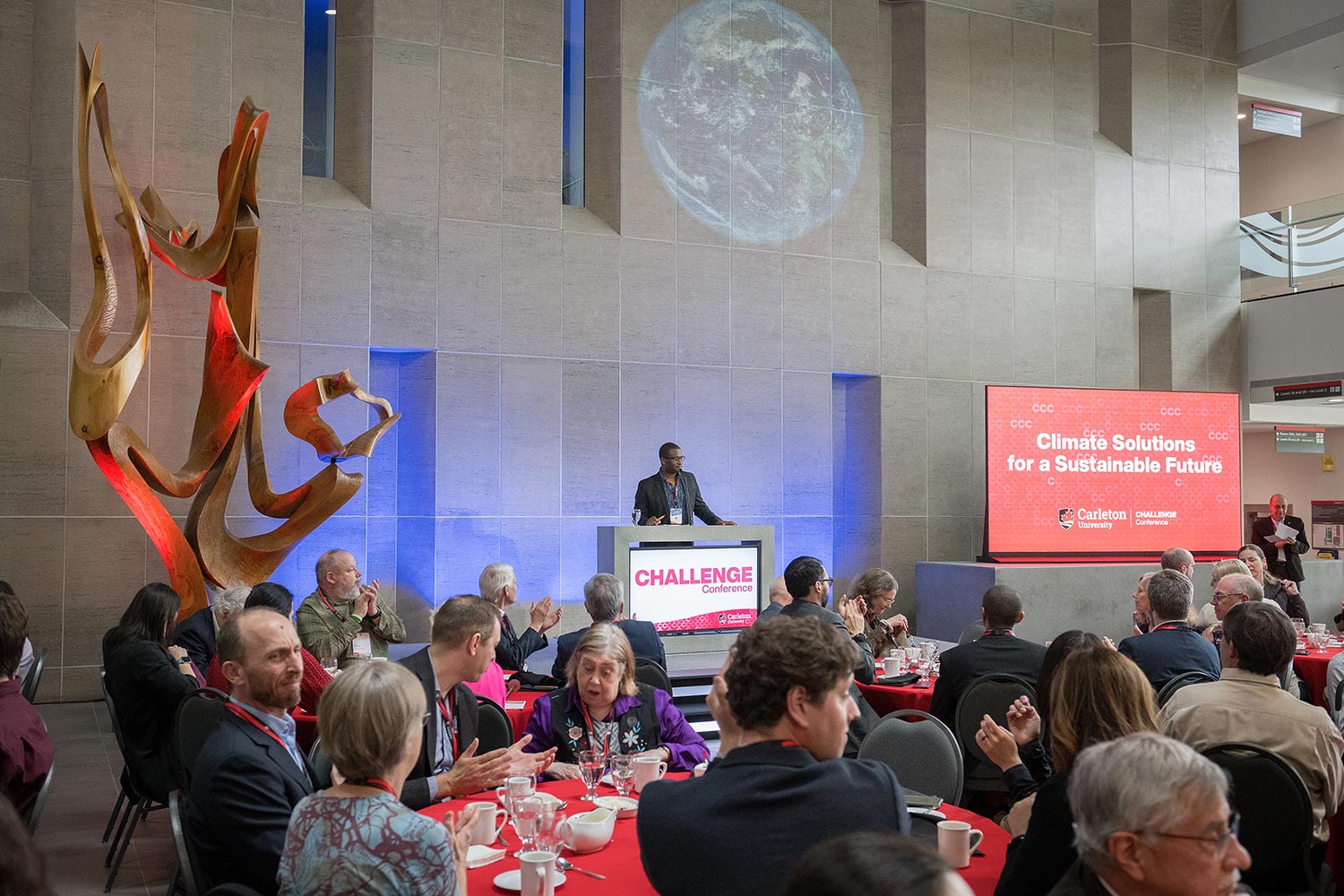 A man addresses a large seated crowd from a podium.