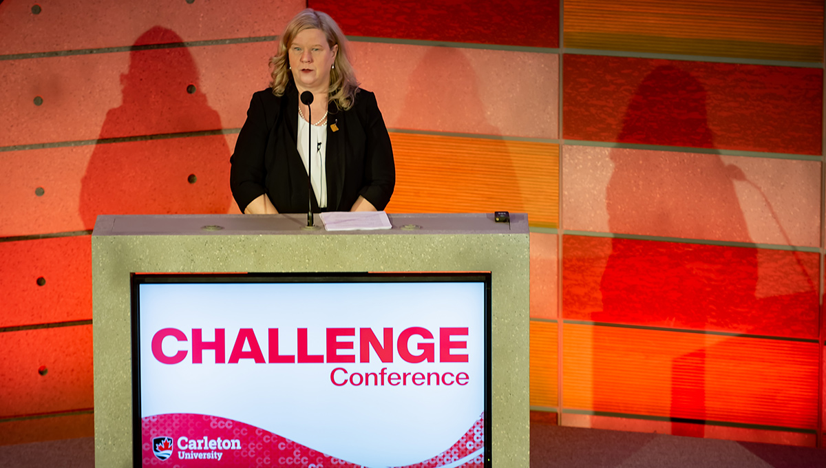 A professionally dressed woman in a black blazer speaks into a microphone from a podium