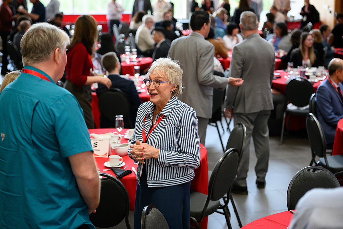 Two people having a conversation at an event, other people can be seen in the background.