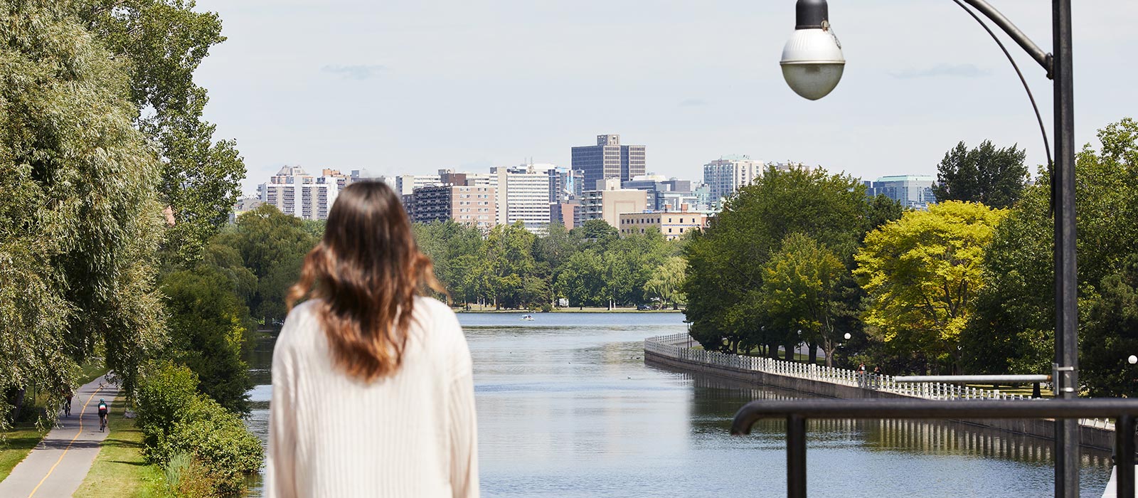 A woman with her back turned looks over a canal with trees along the banks and buildings visible in the distance.