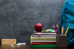 A blackboard eraser, chalk, books, notepads an apple, pencils and backpack sit atop a school desk in front of a classroom chalkboard.