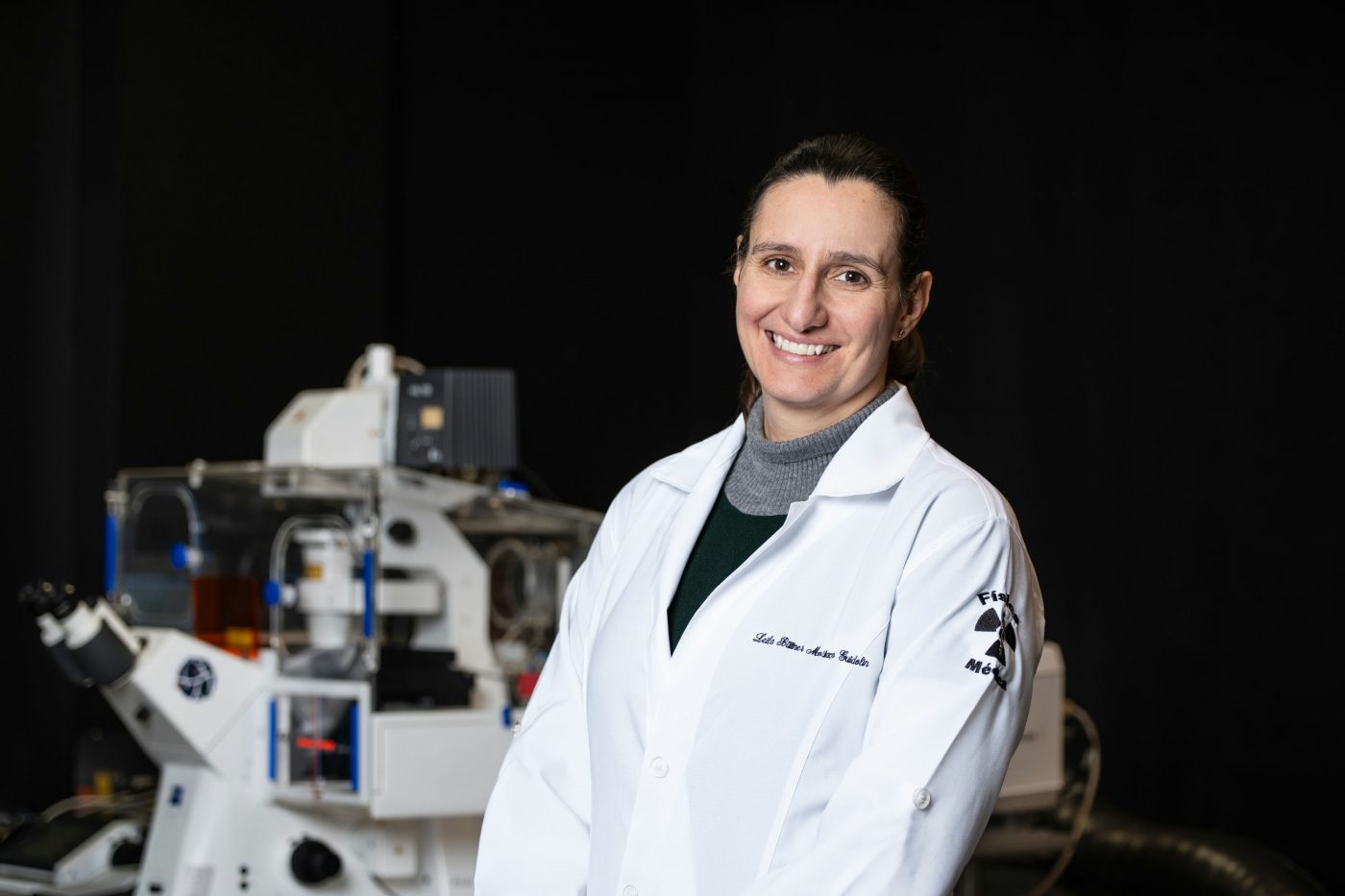 A woman stands in a white lab coat in front of a piece of lab equipment