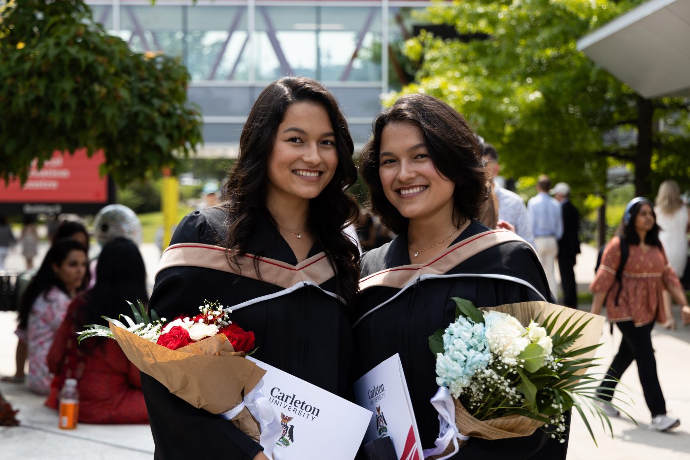 Two graduating students pose with their cu_people_degree and bouquets outside the Athletics Building.