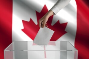 A person's hand puts a ballot in a box while a Canadian flag waves in the background.