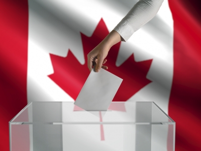 A person's hand puts a ballot in a box while a Canadian flag waves in the background.