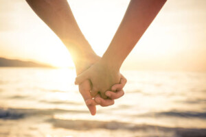 Two people hold hands in front of a seaside sunset.