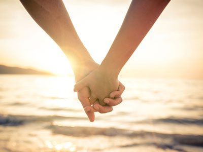 Two people hold hands in front of a seaside sunset.