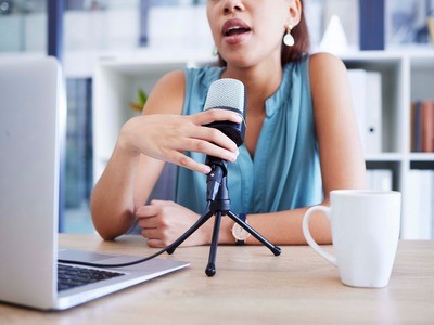 An influencer sits at a desk, speaking into a microphone. A computer is to her left and a mug is to her right side.