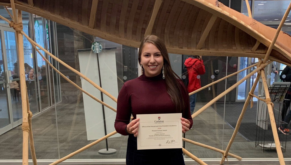 Julianne Dumont holds her Provost's Scholar award while standing in front of a birchbark canoe in MacOdrum Library.
