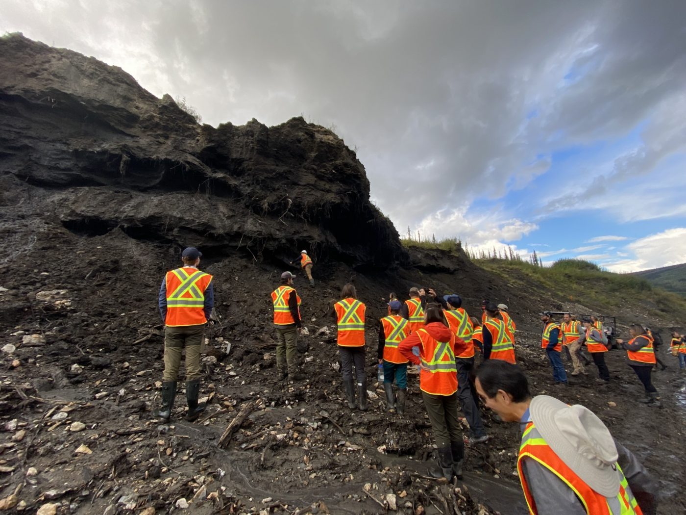 A group of people in orange vests clime a rocky hill.