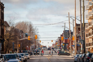 Pedestrians cross a street at a crosswalk in Centretown, Ottawa. Parked cars line the streets.