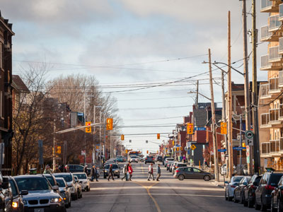 Pedestrians cross a street at a crosswalk in Centretown, Ottawa. Parked cars line the streets.