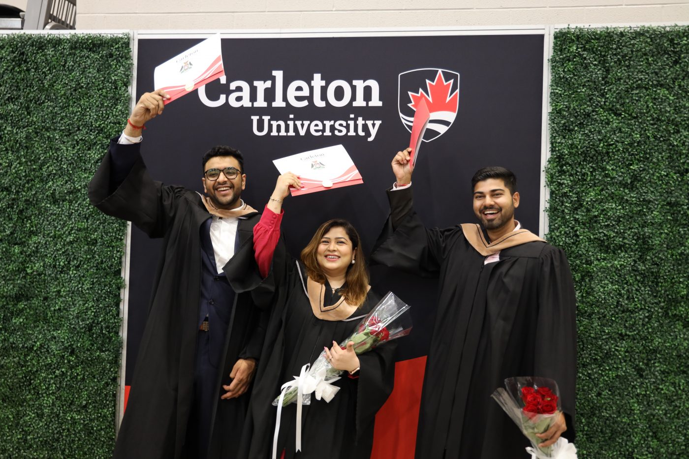 Three graduates pose in front of a Carleton branded banner while holding their cu_people_degree above their heads and smiling