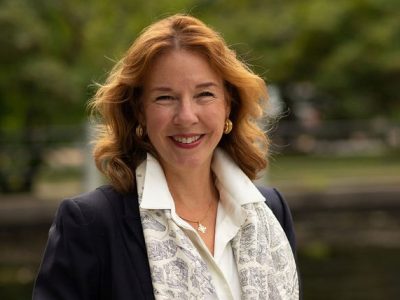 A woman with red hair and a blue dress coat smiles for the camera.