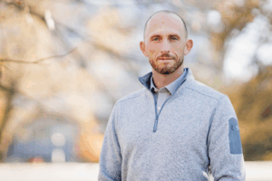 A man in a zip up sweater poses for a photo near a tree.