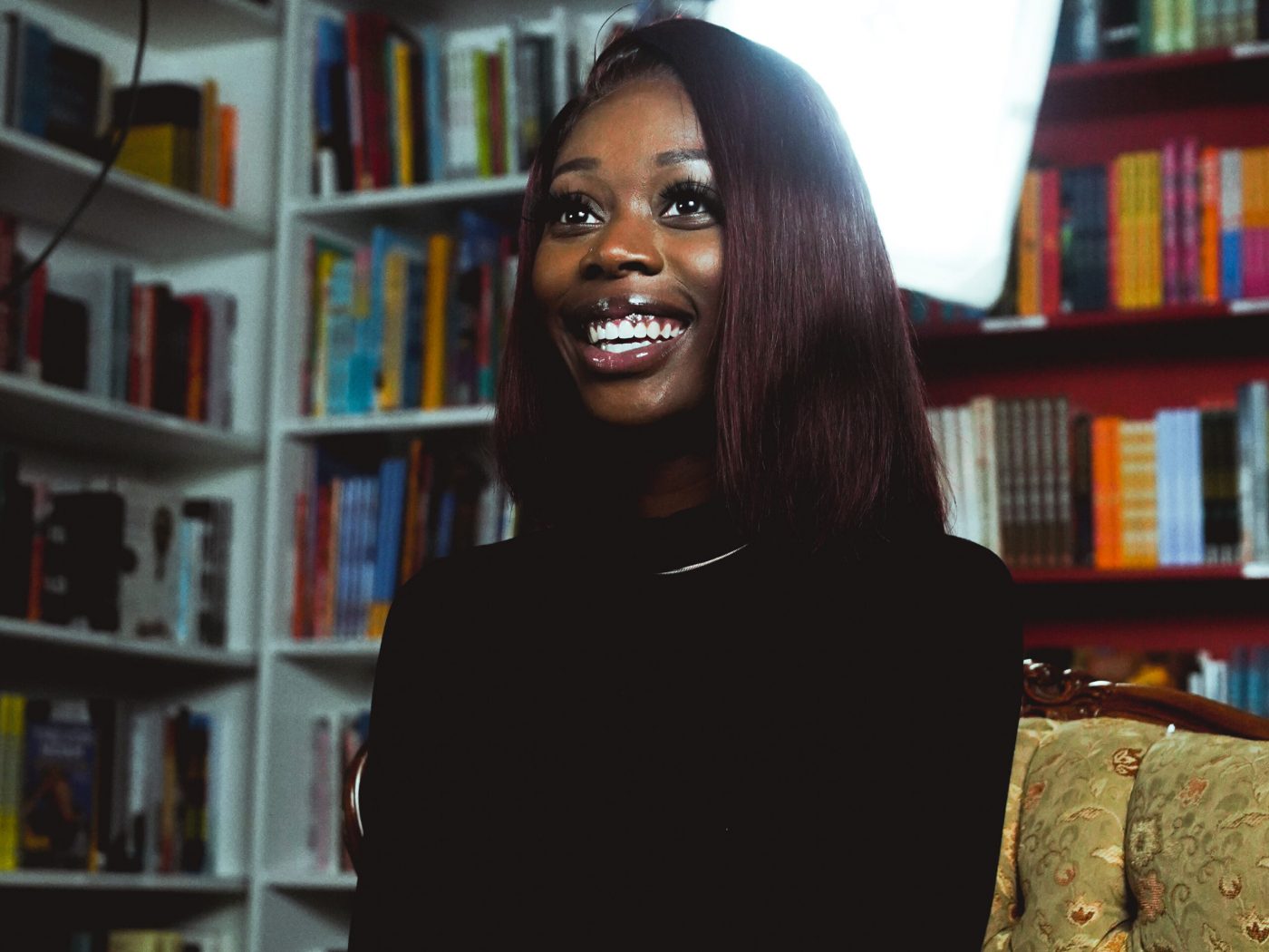 A young woman smiles while sitting in a room surrounded by books. She looks away from the camera.