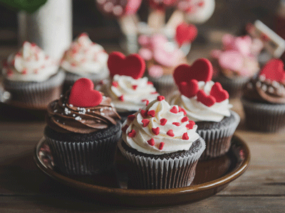 Iced chocolate cupcakes with red candy hearts are set on a brown plate. Additional cupcakes are in the background.