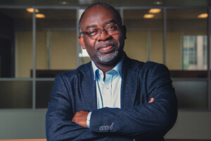 A man with glasses wearing a blue blazer and light blue dress shirt, crosses his arms while looking at the camera