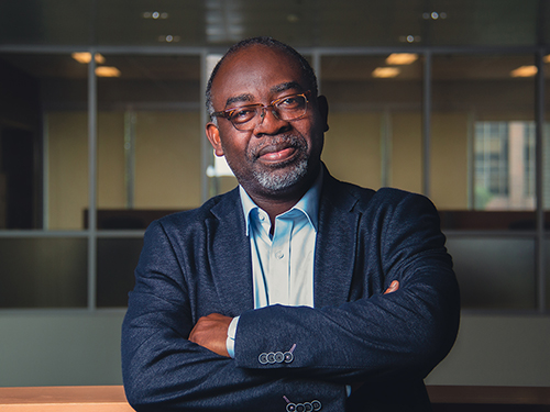 A man with glasses wearing a blue blazer and light blue dress shirt, crosses his arms while looking at the camera