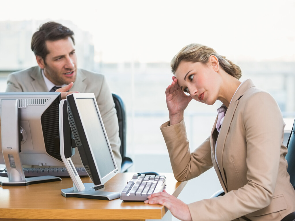 A professional woman sitting in front of a computer appears annoyed by a male co-worker for mansplaining.