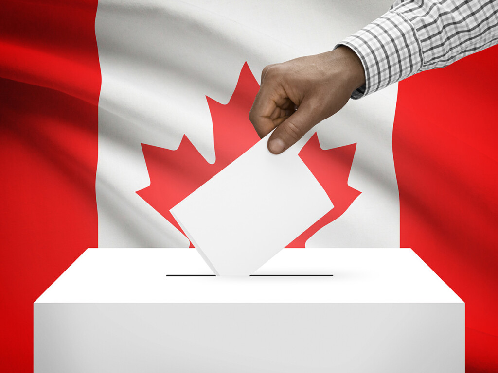 A hand is seen putting a ballot into a ballot box, with a Canadian flag in the background.