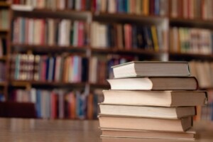 A pile of books sits on a table in a library. Shelves of books are in the background