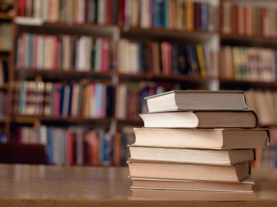 A pile of books sits on a table in a library. Shelves of books are in the background