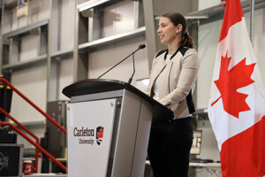 A woman in a business suit speaks at a podium with a Canadian flag visible in the background.