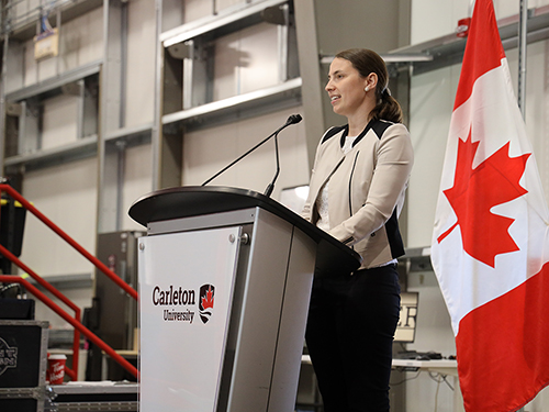 A woman in a business suit speaks at a podium with a Canadian flag visible in the background.