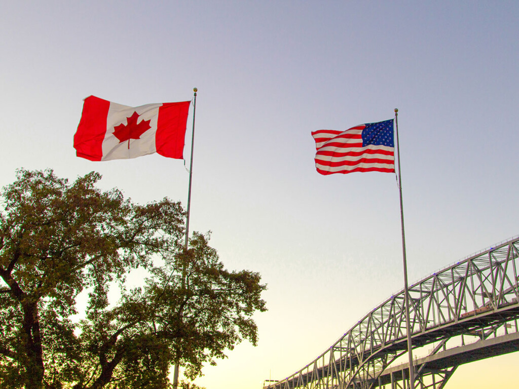 The flags of Canada and the USA with a bridge and trees in the background.