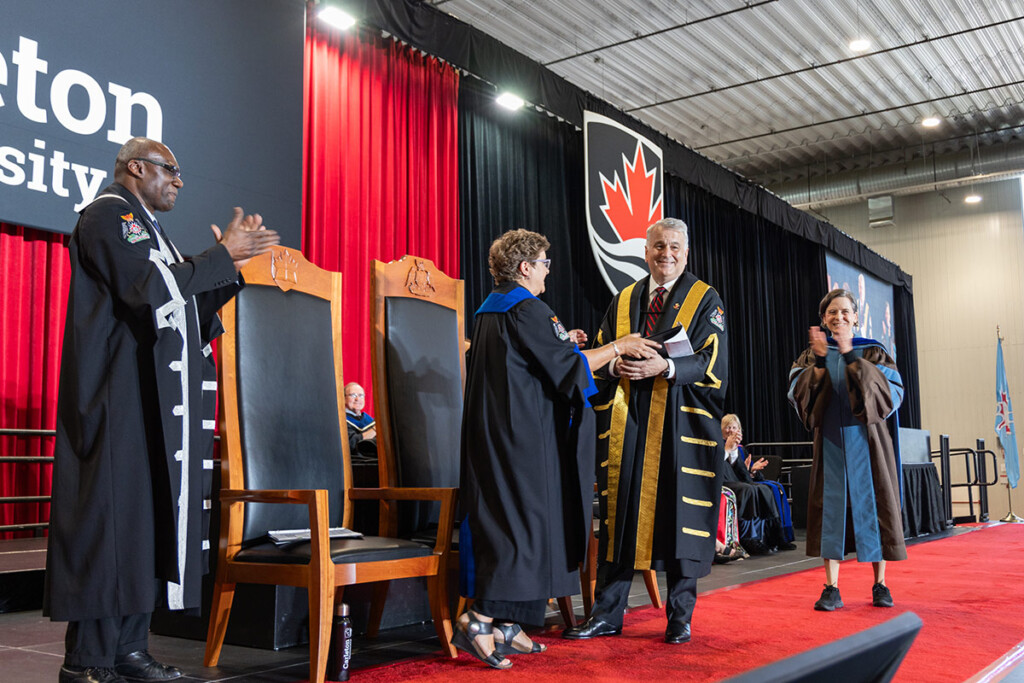 While standing on a large stage, a man wearing academic regalia accepts a document from another person wearing academic regalia.