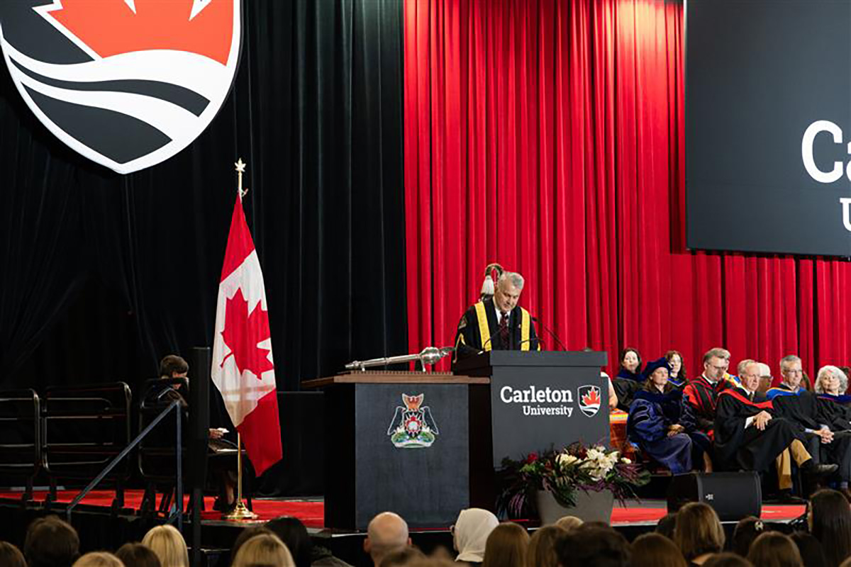 A man stands at a podium on a stage, delivering remarks to the audience seated in front of him.