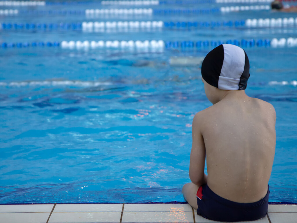 A child swimmer wearing a swim cap sits on the side of a pool (Shutterstock)