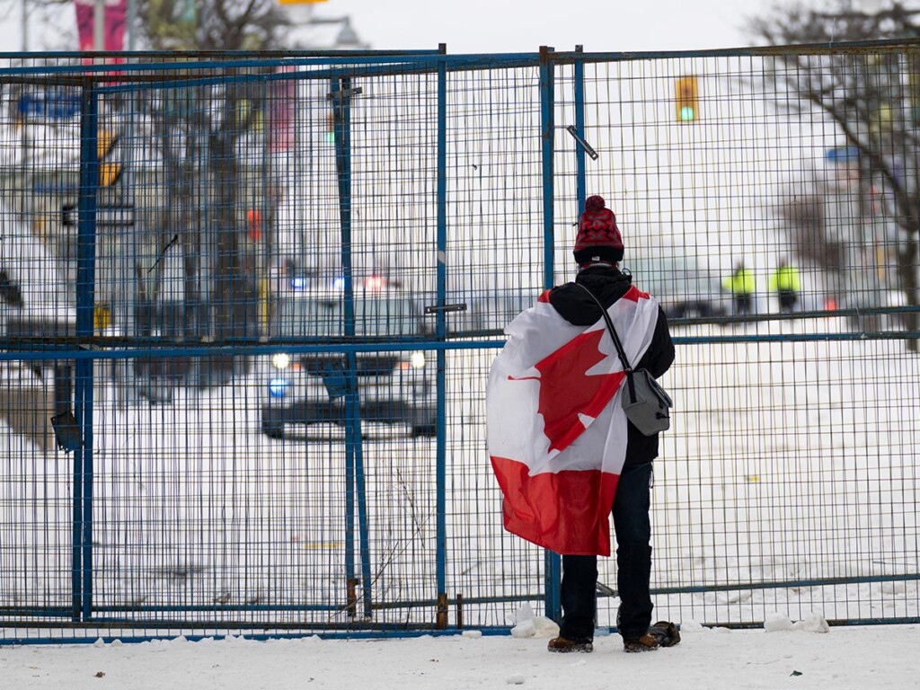 A lone protester stands draped in the Canadian flag at a fence controlling access to streets near Parliament, in Ottawa, Feb. 20, 2022. (Photo: THE CANADIAN PRESS/Adrian Wyld)
