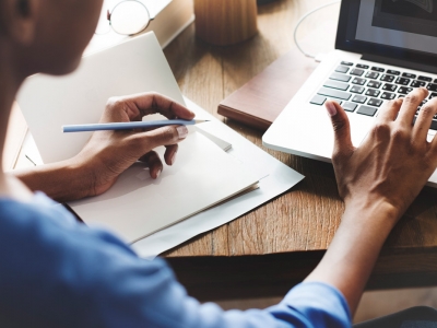 A woman types on a laptop while looking at a notebook.